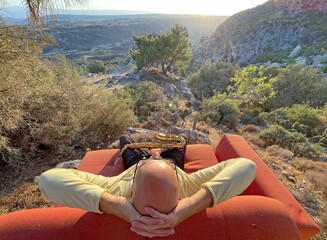 Young stylish bald male saxophonist sits on an orange sofa in mountains of Cyprus in nature, under...