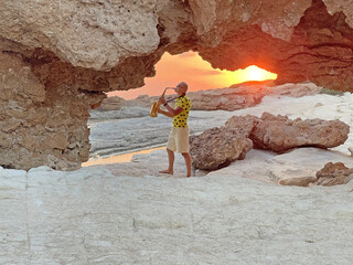 Young stylish bald male saxophonist stands against background of white sea stones, beautiful sunset, sun with saxophone, plays saxophone through cave in mountains. Romantic Musician saxophonist
