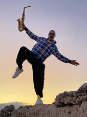 Young stylish bald male saxophonist stands against the background of a beautiful sunset with a saxophone, romantic musician saxophonist, stands on one leg and holds a saxophone, smiling