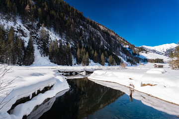 Mountain lake in a snow landscape in front of a mountain forest with needle trees under a blue sky on a sunny bright day in Grossarl Salzburg Austria alps.