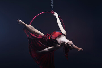 Circus performer woman in red dress doing tricks on red Lyra isolated on black background.