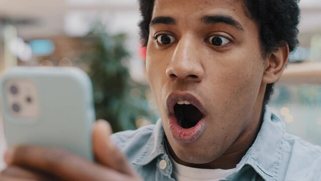 Close-up portrait shocked African American millennial man open mouth in shock looking at smartphone screen feel astonishment scare with bad unexpected news failure fired from work guy looks at phone
