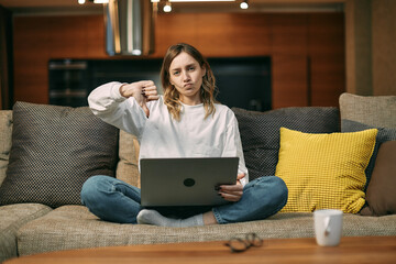 Young girl blogger sit on couch with laptop showing thumb down dissatisfied with services or products. Negative feedback