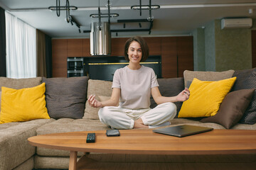 Happy young woman meditate, practice yoga, does mudra gesture, sitting on sofa at home. Healthy lifestyle, stress relief