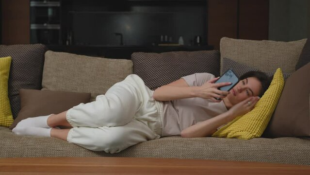 Tired young woman napping on couch, checks social media messages on phone, has a break after hard working day at home