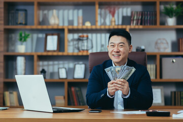 Happy businessman looking at camera and smiling, asian working in office holding american dollars in cash, investing success concept