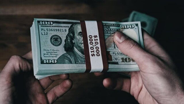 Stack of 10000 American dollars banknotes in male hands over wooden background. Bundles of new design 100 US dollars in currency strap for hundreds. Rich successful businessman holding pile of money.