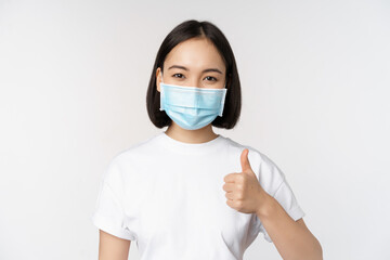 Smiling asian girl in medical mask showing thumbs up, approve something good, praise and compliment...