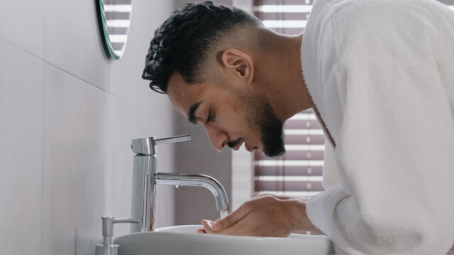 Side view arab spaniard indian arab man guy opens faucet near washbasin at home in hotel bathroom washes face with hot cold water morning beauty ritual male hygiene guy in bathrobe wet facial wash