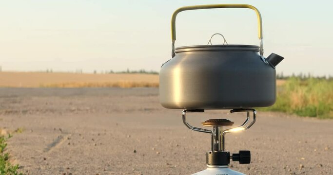 Travel tourist shiny metal kettle pot, boiling hot water in the middle of the road. Outdoor cooking camp equipment on summer sunny background field. Gas propane camping burner. Product packshot shot.
