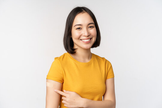 Covid-19 Vaccination Campaign. Smiling Asian Girl Pointing At Her Band Aid On Shoulder, Recommending Get Vaccine From Omicron Coronavirus, White Background