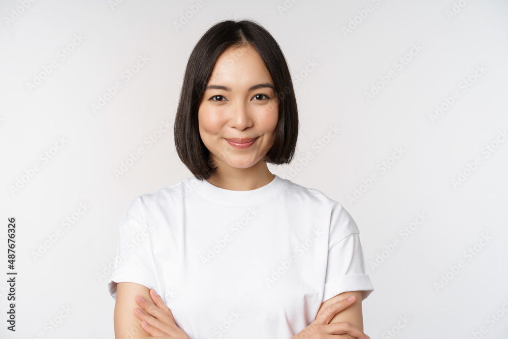 Wall mural Close up portrait of confident korean girl, student looking at camera with pleased smile, arms crossed on chest, standing over white background