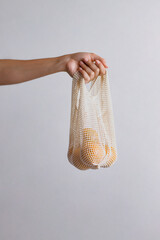 hand holding an ecological fabric bag, with some oranges inside, on a white background