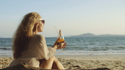 Fototapeta na wymiar Attractive woman sitting in white dress sunglasses, drinking pineapple cocktail Pina Colada. Beautiful beach, sea waves at sunset. Concept rest tropical resort traveling tourism happy summer holidays