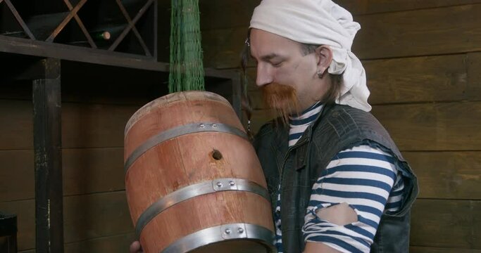 Happy pirate with long ginger mustache and white bandana kisses wooden heavy barrel with rum holding IN arms in ship cabin