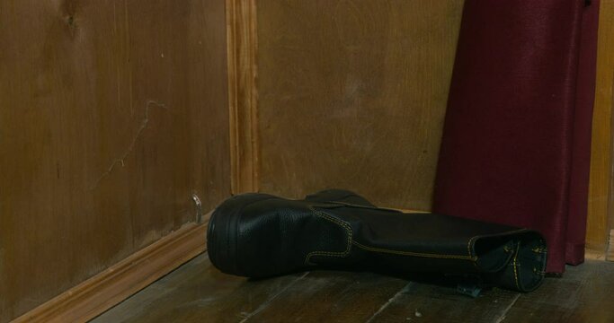 Pirate removes black boot and throws on wooden floor near walls and burgundy curtain IN dark cabin of pirate ship close view