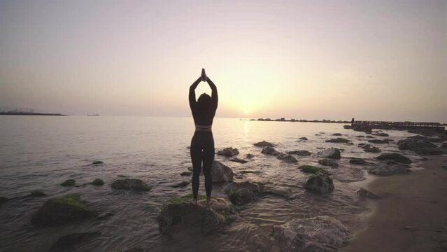 Black afro American young athletic woman practicing yoga at sunrise on a rock in the ocean, morning meditative workout routine