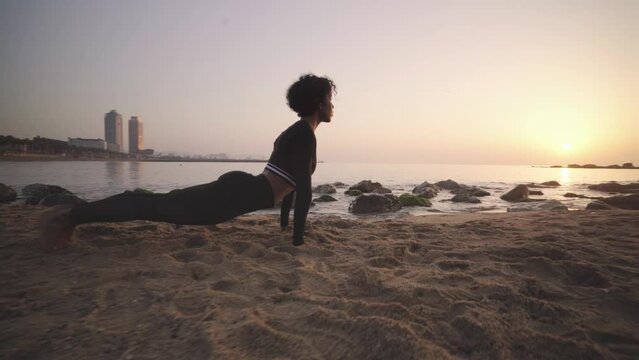 Afro black woman practicing yoga morning routine at sunrise in the sandy beach of Barcelona spain coastline. Healthy shaped young girl fitness workout