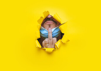 A man in a protective medical mask with a hand gesture in the form of a middle finger (fuck),peers into a torn hole in yellow paper.Protest concept against coronavirus and covid-19 quarantine pandemic