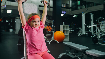 Fit Athletic Baby Girl Work out with Dumbbell, Doing Her Fitness Exercise. Children Activity Training in Modern Gym. Sports Child Workout in Exercising with Barbell, Fitness Center.