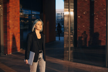 Woman coming out of office building after work, holding laptop in hand and going home in the evening. Businesswoman portrait. Positive person.