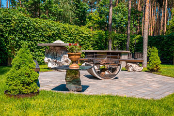 Obraz premium Beautiful sunny backyard with iron fire pit and bench on stone pavement, Garden rest area with fire pit surrounded by green shrubs
