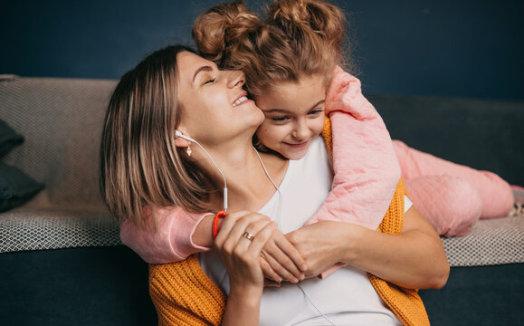 Close-up view of a little girl lying on the couch, hugging her mother's neck from behind, sitting down and listening to music on her headphones. Happy family