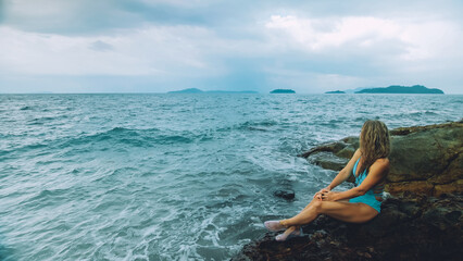 Fototapeta na wymiar Alone attractive woman walk near a rock reef hill in stormy morning rain cloudy sea. Girl in turquoise swimsuit. Concept resort outdoor relax, vacation, loneliness. Dark dramatic view