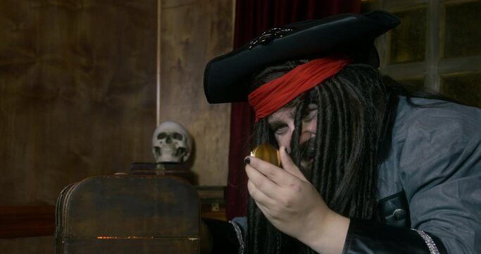 Pirate captain with long hair in cocked hat opens treasure chest to take gold sitting AT table against skull on rack in cabin