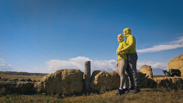 Man and woman in yellow green sportswear. Lovely couple of travelers hug and kiss near old stone enjoying highland landscape. Two travelers are walking against the backdrop of snow-capped mountains. © ivandanru