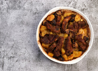 Roasted pork neck with mushrooms and potatoes in a white baking dish on a dark grey background. Top view, flat lay