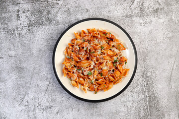 Red chicken pasta with herbs, tomatoes and cheese on a white round plate on a dark gray background. Top view, flat lay