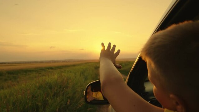 Happy teenager guy rides in a car, waving his hand out the car window. Road travel concept. The boy stretches out his hand from the car window catch the sun glare of the sunset, happy emotions