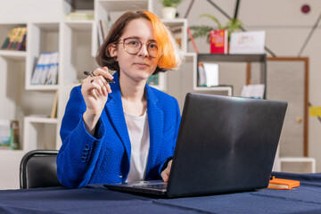 Young modern girl with multi-colored hair in blue business suit sits at table with laptop. He holds pen in his hand and looks into camera.