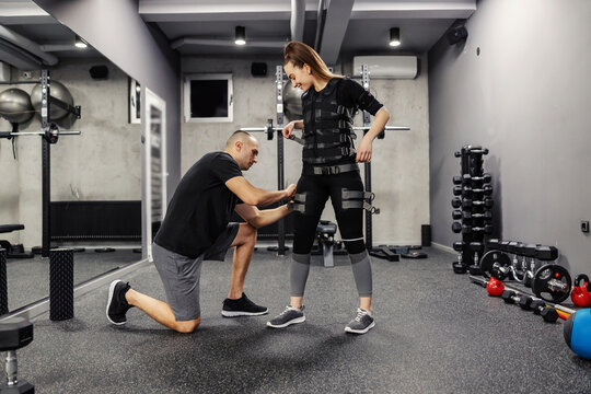 An instructor helps a woman standing to train a suit for EMS technology in a gym.