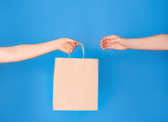 Close-up of cropped hands holding a brown paper takeaway bag isolated on a blue background. The concept of a delivery service. Space for copying. Advertising area