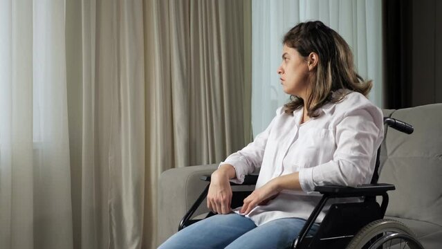 Sad disabled woman with organic damage of central nervous system sits in wheelchair looking out of window behind curtain in living room at home.