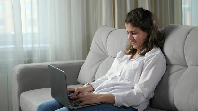 Young disabled woman with cerebral palsy works remotely on computer trying to type text by poorly mobile fingers sitting on sofa by window at home.