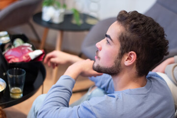 Man sitting on sofa while having get together with friends. Side view of serious Caucasian guy spending time with friends on weekend, eating, talking and watching TV. Party, entertainment concept