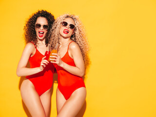 Two beautiful sexy smiling hipster women in red summer swimwear bathing suits.Trendy models with afro curls hairstyle having fun in studio.Hot female isolated on yellow.Drinking lemonade from bottle
