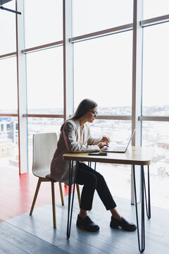 Image of a happy young woman in a jacket smiling and working on a laptop while talking on the phone in a modern office with large windows. Remote work