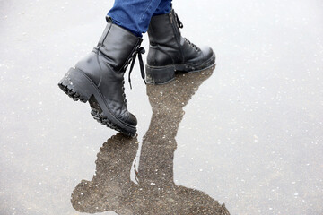 Female legs in black leather boots with reflection in puddle on a street. Rain in city, waterproof footwear for cold weather