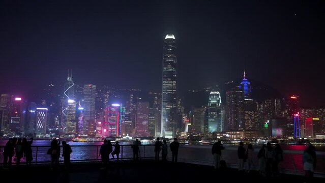 People take photos and enjoy the view of the Victoria Harbour waterfront at nighttime as the Hong Kong Island skyline and skyscrapers are seen in the background.
