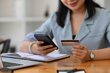Pleased young woman holding credit card and using smart phone for baking online.