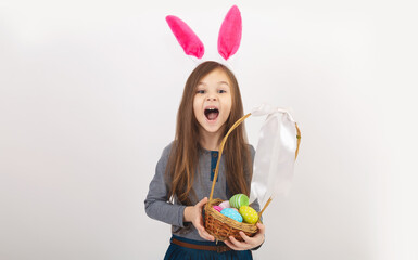 Easter to the child. A cheerful little girl with rabbit ears on her head with a basket of colored eggs in her hands on a white background. Funny crazy happy baby.
