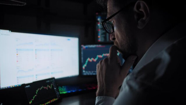 A bearded trader with glasses sitting near to computer monitors and analyzes the stock market date a close-up slow motion