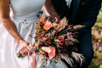 Braut in weißem Kleid hält ihren Brautstrauss in warmen Farben und Gräsern in der Hand