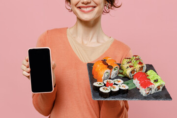 Cropped young woman wear sweater hold makizushi sushi roll served on black plate traditional japanese food use mobile cell phone blank screen workspace area isolated on plain pastel pink background