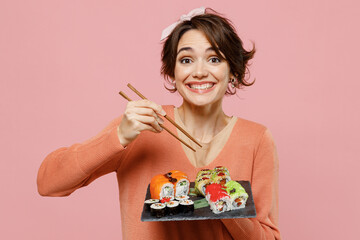 Young satisfied smiling happy woman in casual clothes hold in hand eat with chopsticks makizushi sushi roll served on black plate traditional japanese food isolated on plain pastel pink background