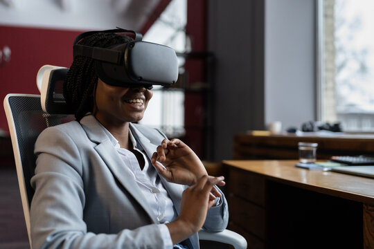 VR training in workplace. Smiling impressed black businesswoman sitting at office table using virtual reality glasses to reduce stress and relax at work. Power of augmented reality AR in business